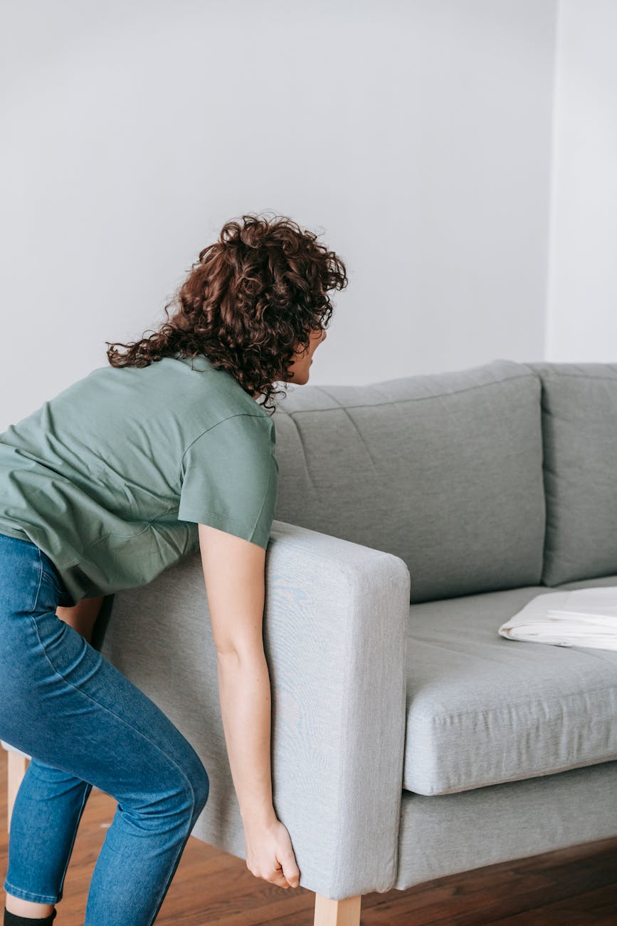 woman in green shirt and blue denim jeans lifting a gray couch